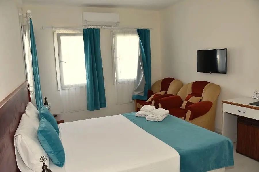 Mavi perdeli, aydınlık bir oda; çift kişilik yatak ve koltuklar